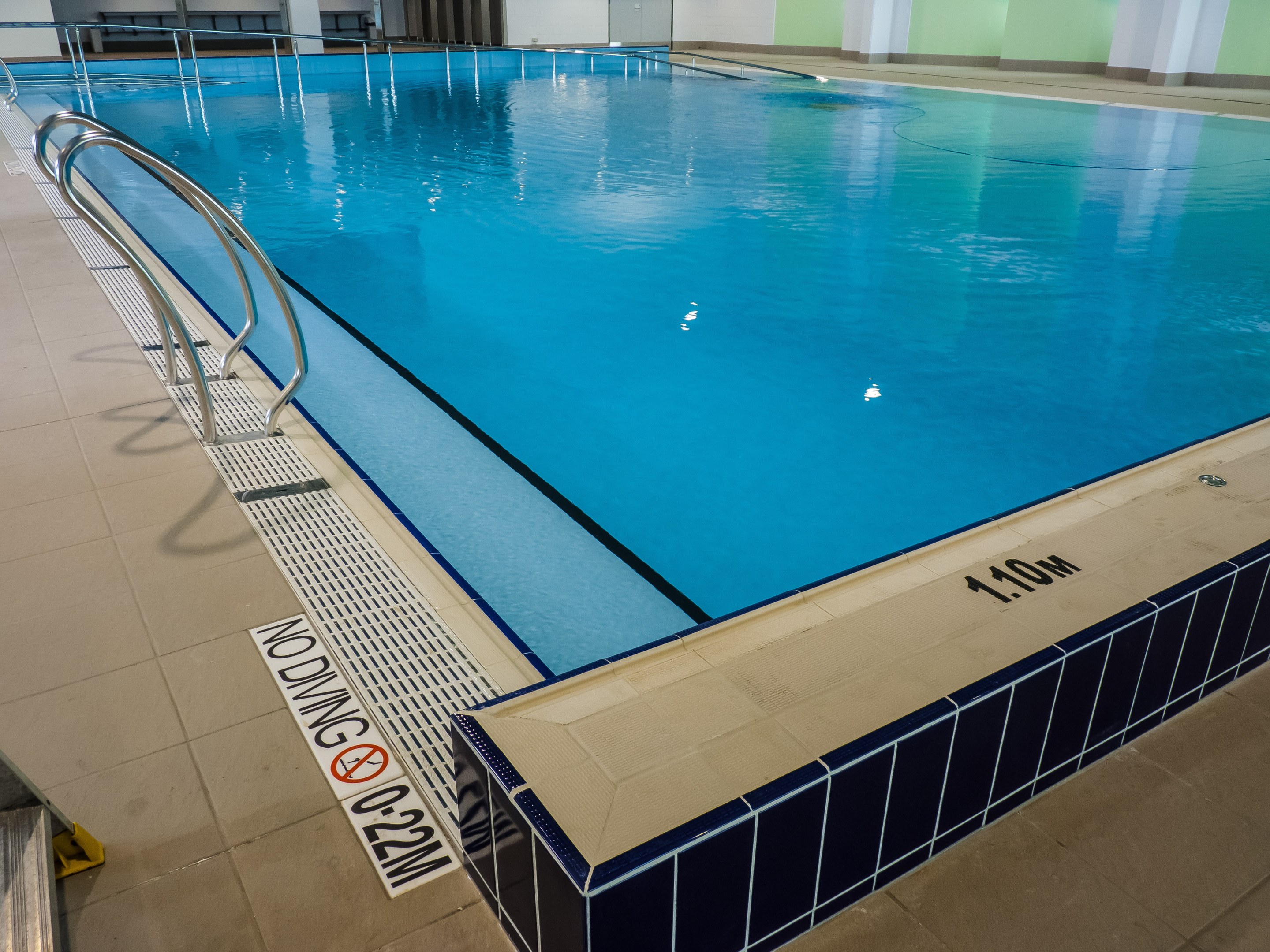 Großes Hallenbad in einem geräumigen Gebäude mit gefliestem Boden, Metallstützstangen, einer Wandtafel, einer Tür und Deckenbeleuchtung.