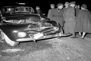 Ein Schwarz-Weiß-Bild eines verbeulten Autos am Straßenrand mit einer Gruppe von Menschen drumherum, die den erheblichen Schaden an der Motorhaße und den zerbrochenen Fenstern begutachten.