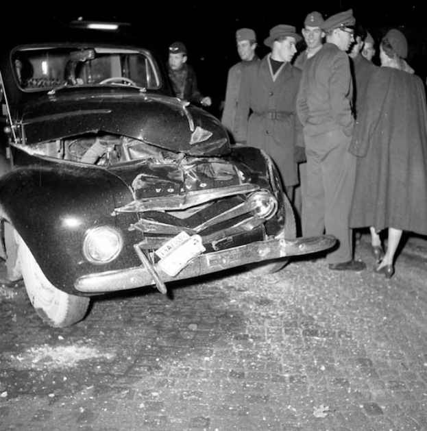 Ein Schwarz-Weiß-Bild eines verbeulten Autos am Straßenrand mit einer Gruppe von Menschen drumherum, die den erheblichen Schaden an der Motorhaße und den zerbrochenen Fenstern begutachten.