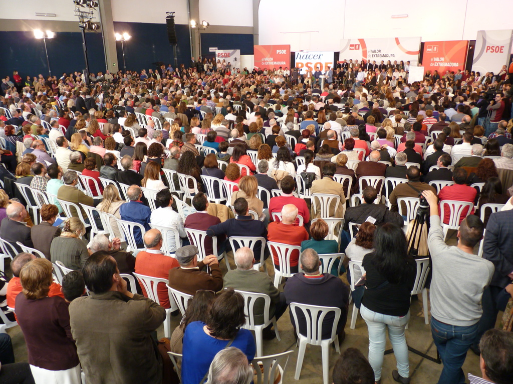 Ein volles Stadion mit vielen sitzenden und stehenden Menschen, eine Bühne mit zwei Rednern an einem Podium und mehreren Bannern und Lichtern in der Nähe.