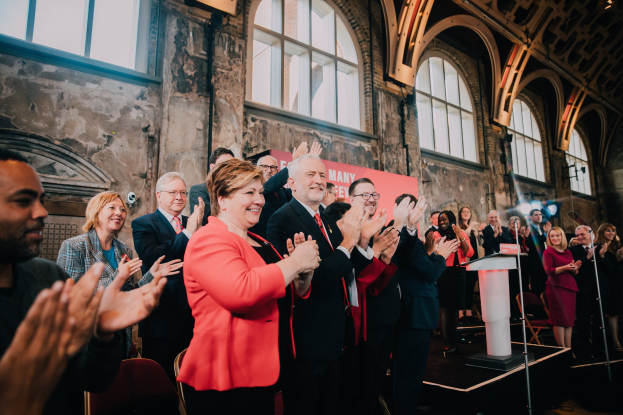 Eine Gruppe von Menschen, die vor einem Publikum klatscht, mit einem Podium, einem Mikrofon, Stühlen, einer Wand, einem Banner mit der Aufschrift "Covid-19 New York City Mayor Bill de Blasio" und Fenstern im Hintergrund.