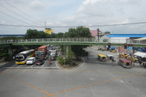 Eine belebte Straßenszene mit verschiedenen Fahrzeugen, Fußgängern, einer Brücke, Ampeln, Gebäuden, Bäumen und Strommasten unter einem bewölkten Himmel.