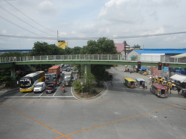 Eine belebte Straßenszene mit verschiedenen Fahrzeugen, Fußgängern, einer Brücke, Ampeln, Gebäuden, Bäumen und Strommasten unter einem bewölkten Himmel.