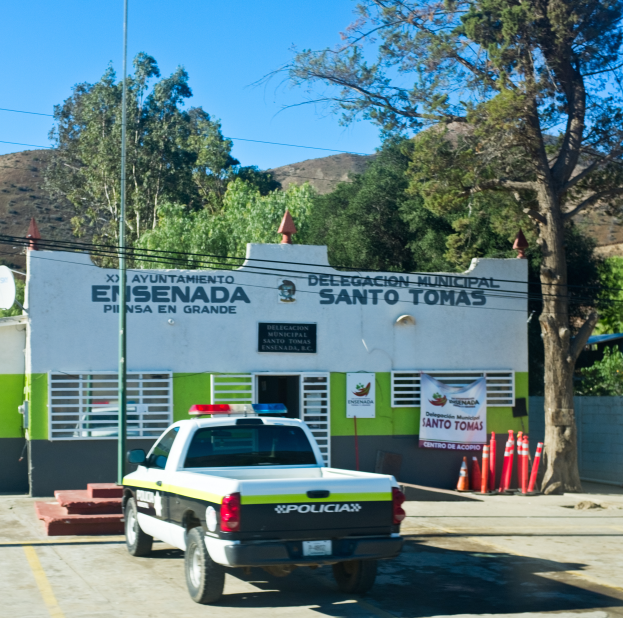 Ein Polizeiwagen vor einem Gebäude mit einem Schild, Fenstern und einer Tür geparkt, mit einem Banner an der Wand, Verkehrsampeln, einem Straßenschild, einer Satellitenschüssel, Kabeln, Bäumen, Hügeln und einem bewölkten Himmel im Hintergrund.