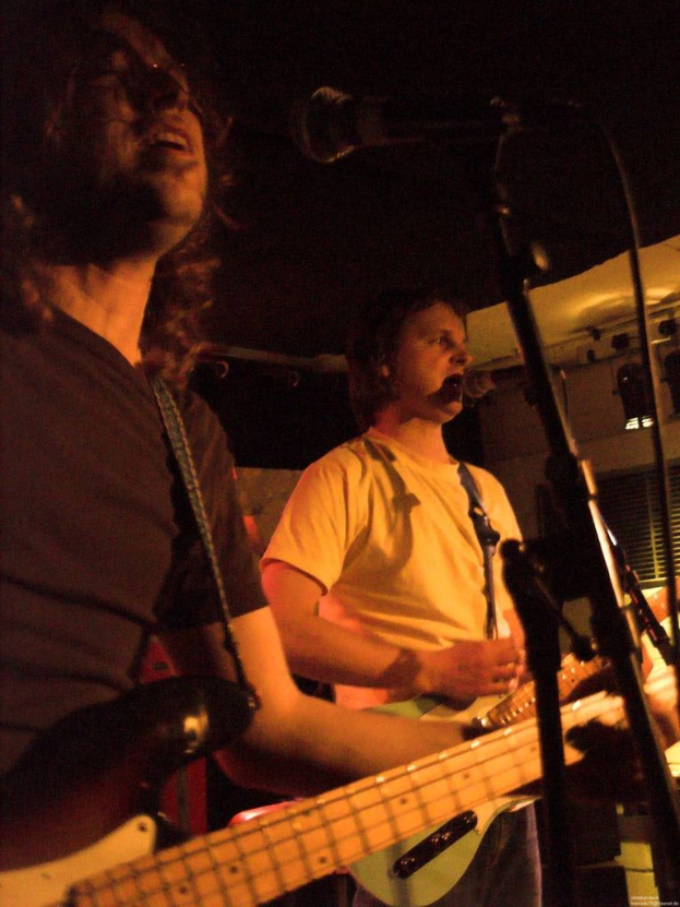 Eine Person in einem weißen Hemd singt in ein Mikrofon, während sie eine grüne Gitarre spielt und neben einer anderen Person in einem schwarzen Hemd, Brille und einer schwarzen Gitarre steht, mit Scheinwerfern und Musikinstrumenten im Hintergrund.