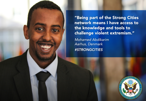 Ein Mann im Anzug und Krawatte lächelt in die Kamera mit einem Logo und Text auf der rechten Seite, der besagt "Teil des starken Städte-Netzwerks zu sein bedeutet, Zugang zu Wissen und Werkzeugen zu haben, um gewalttätigen Extremismus in Frage zu stellen", mit einem unscharfen Hintergrund.