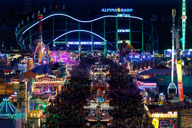 Eine große Menschenmenge umringt eine hell erleuchtete Karnevalsszene in der Nacht, umgeben von Vergnügungsgeräten, Pfählen, Fahnen und Namensschildern, mit einem dunklen Hintergrund.