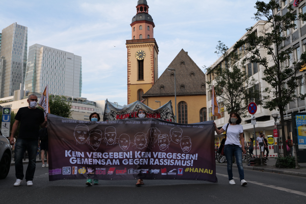 Eine Gruppe von Menschen mit Masken, die eine Straße entlang marschieren und eine Fahne mit Text und Bildern halten, mit einem geparkten Auto auf der linken Seite, Gebäuden, Bäumen, Schildern, Polen, einem Uhrenturm und einem klaren blauen Himmel im Hintergrund.
