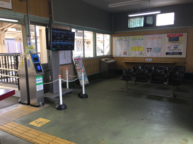 Ein belebter Bahnhofseingang mit Reihen von Stühlen, einer Service-Theke, Ticketautomaten, Kettenzäunen, Informationsanzeigen, Wandplakaten, Deckenbeleuchtung und Geländern.