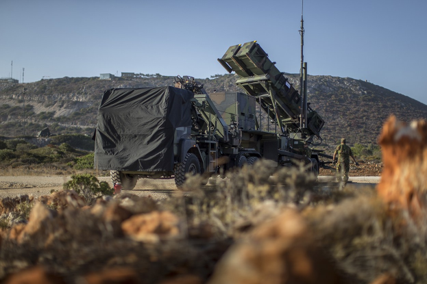 Militärlastwagen mit einer Rakete auf der Rückseite, der entlang einer Schotterstraße fährt, umgeben von Vegetation und Felsen, mit Hügeln, Bäumen, Gebäuden, Strommasten und Himmel im Hintergrund.