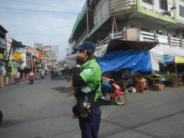 Ein Polizeibeamter in grüner und blauer Uniform und blauer Mütze steht an der Seite einer Straße, spricht auf einem Handy, mit Fahrzeugen, die hinter ihm auf der Straße fahren, Ständen mit verschiedenen Gegenständen zu seiner Rechten und einem bewölkten Himmel über einem Gebäude mit Fenstern im Hintergrund.