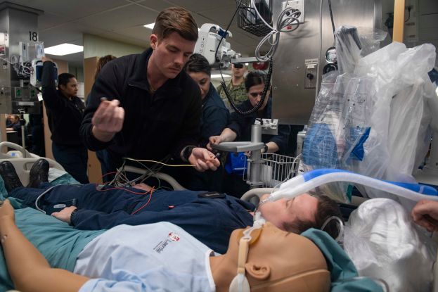 Ein Mann liegt auf einem Krankenhausbett neben einer Puppe, umgeben von einer Gruppe von Menschen und medizinischen Geräten, in einem hell erleuchteten Raum, wahrscheinlich während eines Nationalgarde-Programms für Ersthelfer.