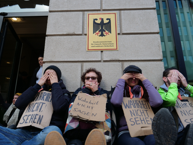 Eine Gruppe von Menschen sitzt vor einem Gebäude, hält Schilder hoch, einige verdecken ihre Gesichter, mit einem Logo und einer Tafel an der Wand und einer Glastür auf der rechten Seite.