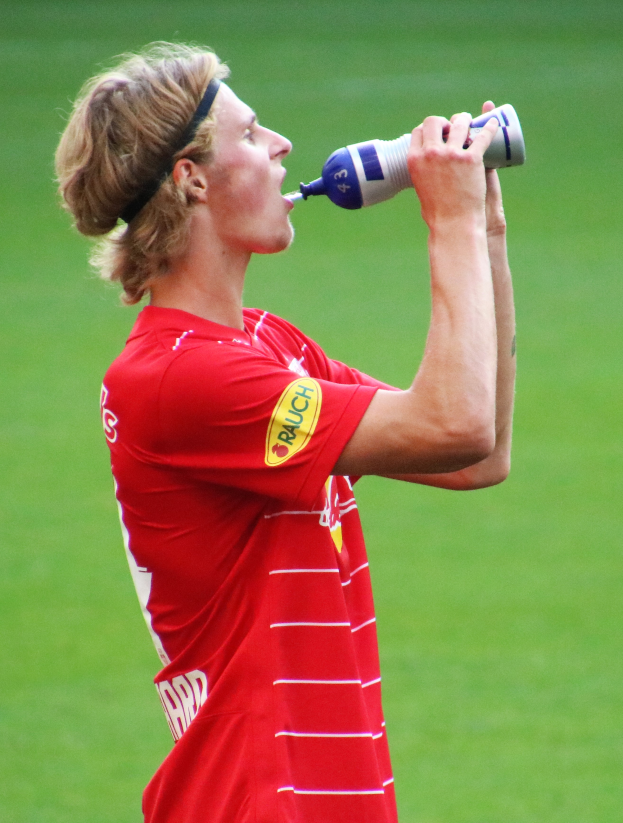 Mann in rotem T-Shirt trinkt aus einer Flasche auf einem Fussballfeld.