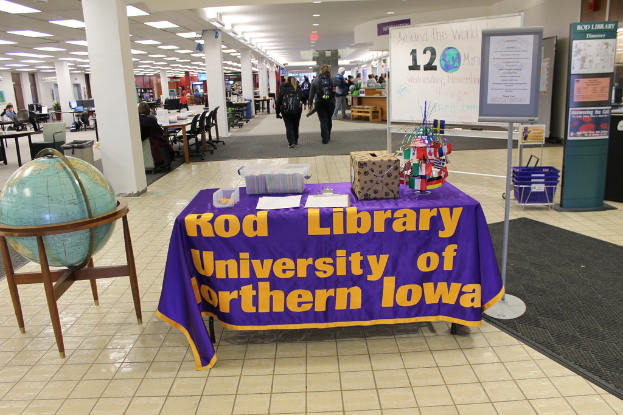 Eine Expo-Szene mit einem Tisch, auf dem verschiedene Landesflaggen, eine Box, Papiere und ein Banner sowie links daneben eine Weltkugel, rechts daneben eine Anschlagtafel mit einigen Menschen in der Nähe und mehrere Tische und Stühle im Hintergrund zu sehen sind.