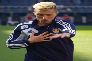 Ein junger Mann in einem blauen T-Shirt steht auf einem Fußballfeld und hat die Arme ausgestreckt und einen entschlossenen Gesichtsausdruck, mit einem unscharfen Hintergrund.