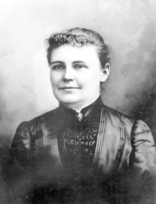 Ein Schwarz-Weiß-Foto einer Frau in einem Kleid, die mit gefalteten Händen und erhobenem Haupt steht und Entschlossenheit und Stärke ausstrahlt.