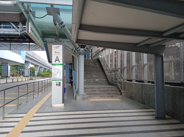 Ein Bahnsteig mit Treppe, eine Straße mit Geländer auf der linken Seite und Hintergrund-Elemente einschließlich Bäume, Pfähle, Gebäude und eine Brücke, mit Rohren und einem Schild am oberen Ende des Bahnsteigs.