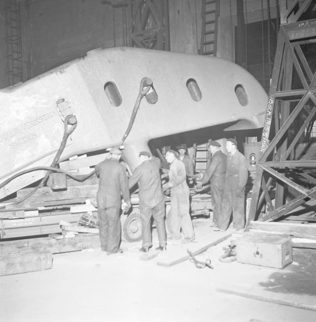 Eine Gruppe von Männern in Mützen arbeitet an einem Flugzeug in einer Schwarz-Weiß-Fabrikumgebung, umgeben von Metallobjekten, Leitern und anderen Gegenständen.