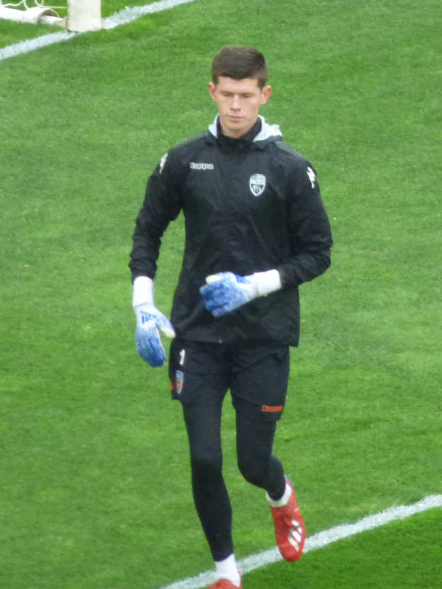 Ein Mann in einer schwarzen Jacke und blauen Handschuhen geht auf einem Fußballfeld, mit einem Torpfosten im Hintergrund.