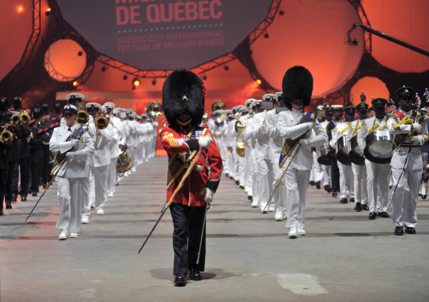 Eine Gruppe uniformierter Personen marschiert die Straße entlang, einige spielen Musikinstrumente, mit einem beleuchteten Schild im Hintergrund, das die Eröffnungszeremonie des Montreal International Festivals of Military Bands anzeigt.