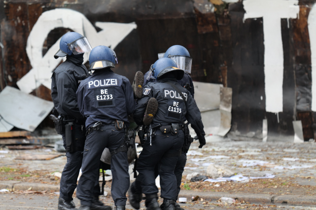 Polizisten in schwarzen Uniformen und blauen Helmen stehen vor einem mit Graffiti bedeckten Gebäude während einer Straßenschlacht, mit verstreuten Papieren und Gegenständen auf dem Boden.