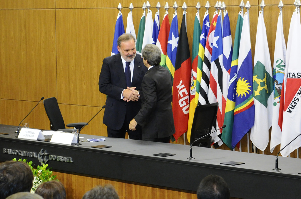 Zwei Personen geben sich die Hand hinter einem Tisch mit Mikrofonen, Namensschildern, Gläsern und Papieren, mit Flaggen dahinter und einem Publikum davor.