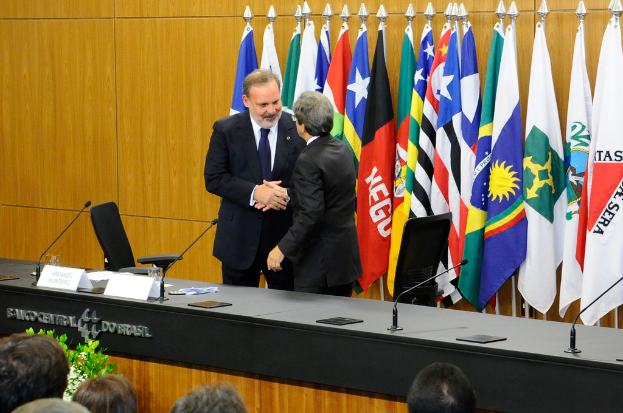 Zwei Personen geben sich die Hand hinter einem Tisch mit Mikrofonen, Namensschildern, Gläsern und Papieren, mit Flaggen dahinter und einem Publikum davor.