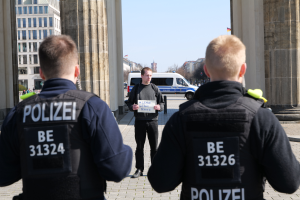 Drei Polizeibeamte stehen vor einem Gebäude, wobei ein Mann in der Mitte ein Blatt Papier hölt, flankiert von Säulen, mit Fahrzeugen, Menschen, Bäumen, Gebäuden und Himmel im Hintergrund.