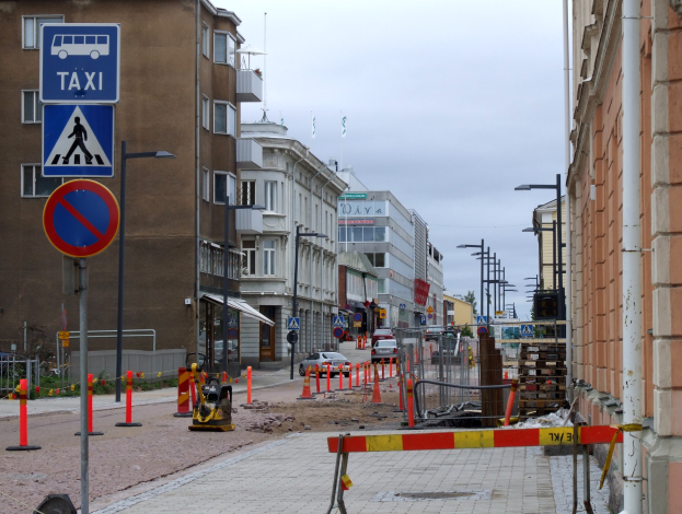 Stadtstraße mit Gebäuden, Straßeninfrastruktur, Kraftfahrzeugen, einer Baustelle, Bäumen und einem bewölkten Himmel.