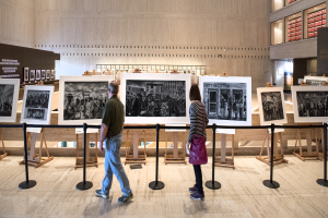 Ein Mann in einem grünen T-Shirt und blauen Jeans und eine Frau mit einer pinken Tasche gehen durch ein Museum mit cremefarbenen Wänden und sehen sich Fotos in mehreren Rahmen an.