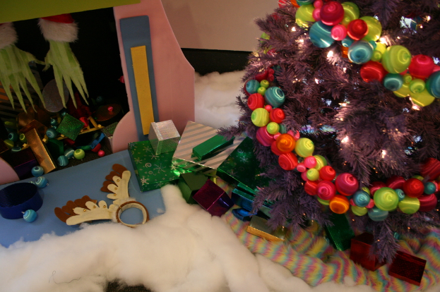 Drinnen steht rechts ein geschmückter Weihnachtsbaum mit Lichterketten und Schmuck, in der Mitte Geschenkboxen und Dekorationen, in der linken oberen Ecke eine Hütte mit Dekorationen und unten Wolle und Baumwolle.