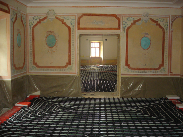 Ein Raum mit einer Fußbodenheizung in der Mitte, umgeben von Wänden und einem Fenster im Hintergrund, mit verschiedenen Gegenständen auf dem Boden, die mit dem Heizsystem in Zusammenhang stehen.