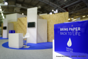 Ein Messestand mit einem blauen und weißen Schild, auf dem 'Bring Paper Back to Life' steht, auf der rechten Seite, mit unscharfen Stühlen, Tischen, einem Fernseher und Deckenleuchten im Hintergrund.
