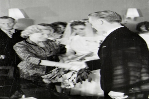 Ein Schwarz-Weiß-Foto einer Braut und eines Bräutigams beim Austausch der Trauringe, wobei die Braut ein weißes Kleid und eine Schleier trägt und einen Strauß hält, und der Bräutigam einen Anzug und eine Krawatte trägt, mit Wandlampen im Hintergrund.