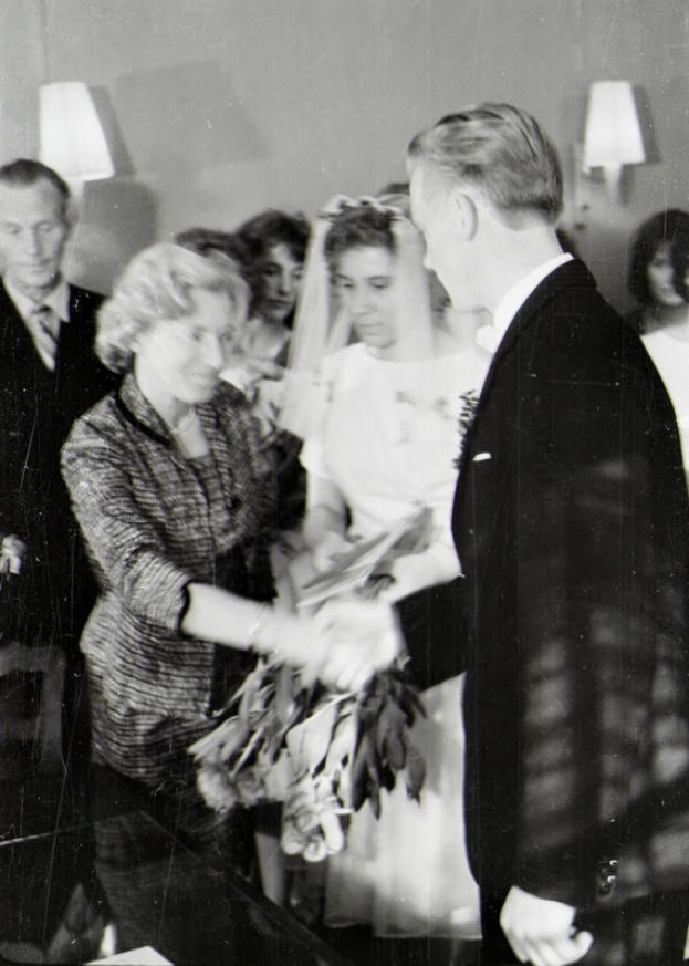 Ein Schwarz-Weiß-Foto einer Braut und eines Bräutigams beim Austausch der Trauringe, wobei die Braut ein weißes Kleid und eine Schleier trägt und einen Strauß hält, und der Bräutigam einen Anzug und eine Krawatte trägt, mit Wandlampen im Hintergrund.