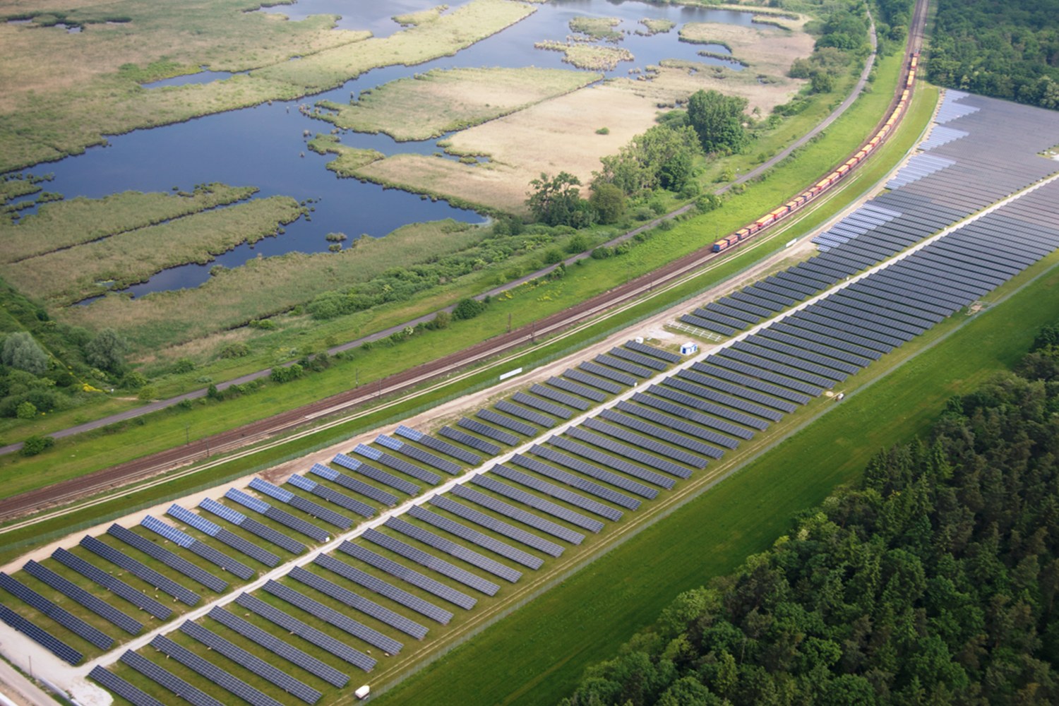 Luftaufnahme einer Solarpark mit Panelen, umgeben von Bumen, Gras, Wasser und einer Eisenbahnschiene in der Nhe.