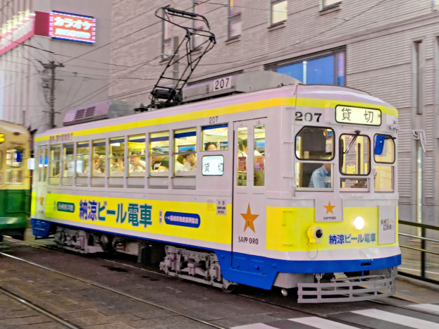 Eine gelbe und blaue Straßenbahn auf einer Stadtstraße in der Nacht mit Menschen darin, Gebäuden mit Fenstern, Strommasten mit Drähten und Schildern im Hintergrund.