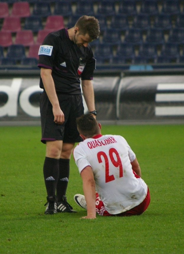 Ein Fussballspieler in Sportkleidung sitzt neben einem Schiedsrichter in einer Stadionkulisse mit Plakaten und Stühlen im Hintergrund.