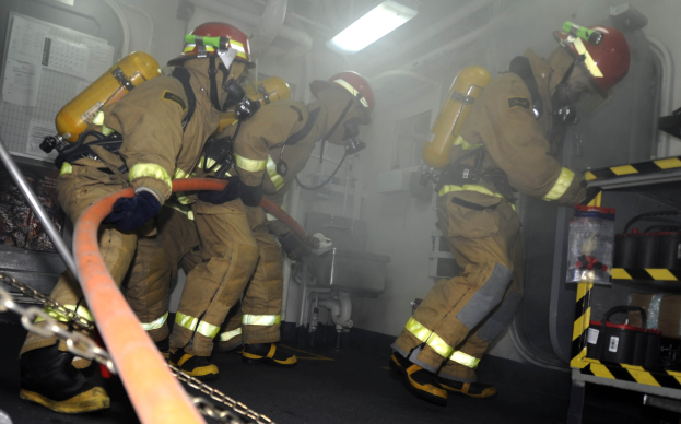 Feuerwehrleute in Schutzausrüstung arbeiten an einem Löschfahrzeug, mit Equipment-Racks und industriellem Hintergrund.