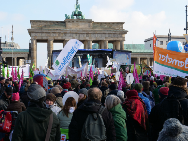 Eine große Menschenmenge steht vor dem Brandenburger Tor in Berlin, Deutschland, einige tragen Mützen und halten Taschen oder Schilder, eine Bühne und Laternenmasten sind im Hintergrund unter einem bewölkten Himmel zu sehen.