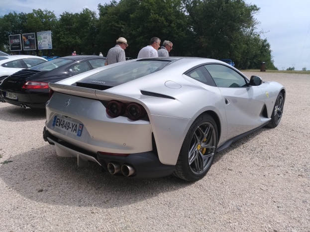 Roter Ferrari 812 Superfast auf dem Goodwood Festival of Speed, umgeben von Zuschauern mit Mützen, Bäumen, Informationsschildern und einem bewölkten Himmel im Hintergrund.