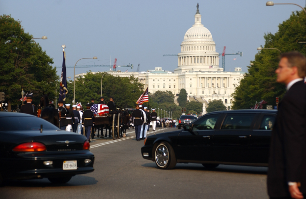 Trauerzug mit Menschen in Mützen und Fahnen vor dem Kapitolsgebäude, Fahrzeuge auf der Straße, Laternenpfähle, Bäume und Gebäude mit Fenstern im Hintergrund unter einem klaren Himmel.