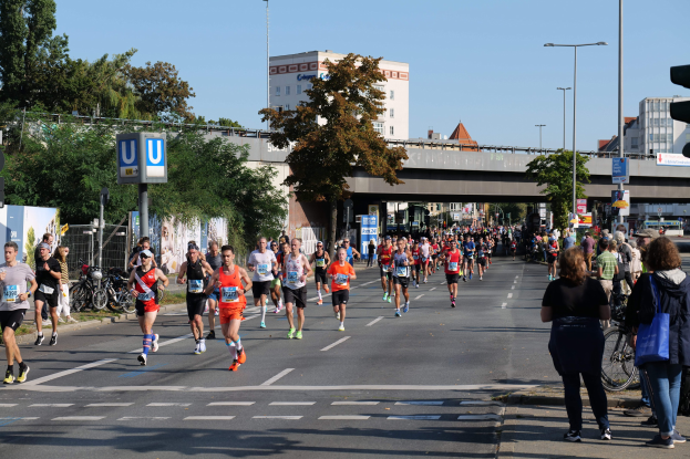 Gruppe von Menschen, die auf einer baumbestandenen Straße mit Laternen, Schildern, Fahrrädern, einem Zaun, Gras, einer Brücke, Gebäuden und einem klaren blauen Himmel laufen.