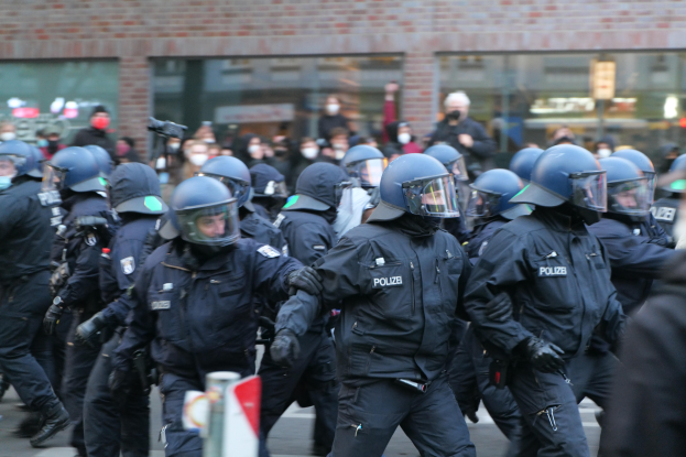 Eine Gruppe von Polizisten in Einsatzausrüstung, einschließlich Helmen und Handschuhen, geht eine Straße entlang mit einem unscharfen Hintergrund, der ein Gebäude mit Glaswänden und eine Stange mit einem Schild zeigt.