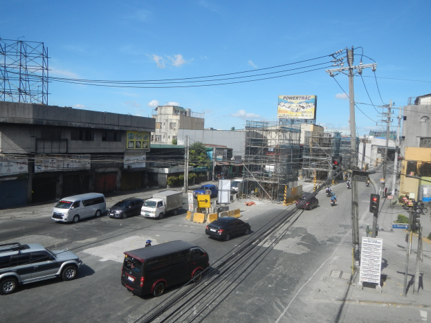 Eine belebte Stadtstraße mit hohen Gebäuden, mehreren Fahrzeugen, Strommasten mit Drähten, Ampeln, Schildern, Bäumen und einem bewölkten Himmel.