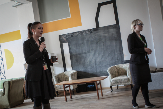 Zwei Frauen, eine hält ein Mikrofon, mit einer Couchgarnitur, einem Tisch und einer Wand im Hintergrund.
