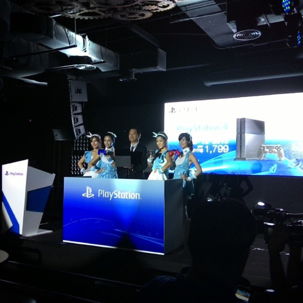 Menschen auf einer Bühne in der Nähe eines Podiums mit 'Playstation' darauf, mit einem Projektionsschirm an der Wand dahinter.