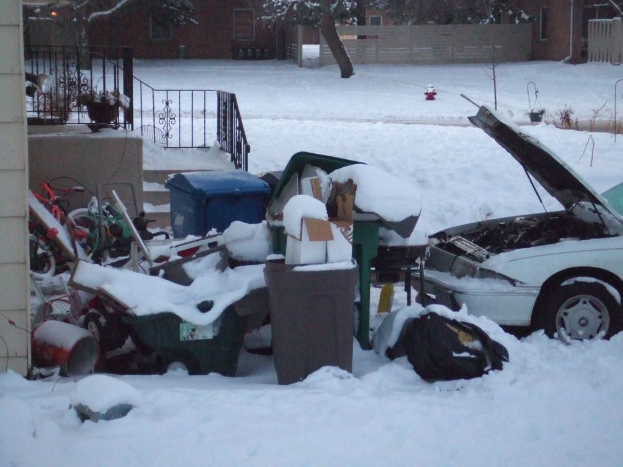 Eine Außenaufnahme mit Müll in der Mitte, einem Auto auf der rechten Seite, einer Treppe auf der linken Seite, Bäumen oben und Eis am Boden.
