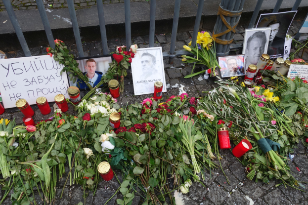 Ein Denkmal in Kiew, Ukraine, mit Blumensträußen, Kerzen, Foto-Rahmen und anderen Gegenständen, die vor einem Zaun auf dem Boden plaziert sind, mit einer Straße im Hintergrund.